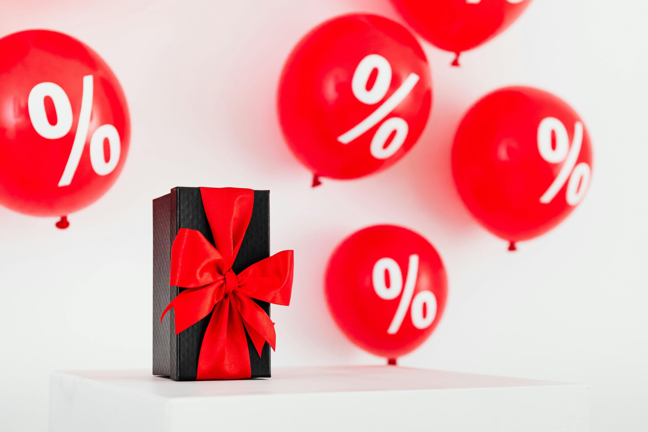 Stylish black gift box with red ribbon surrounded by red percentage balloons on a white background.
