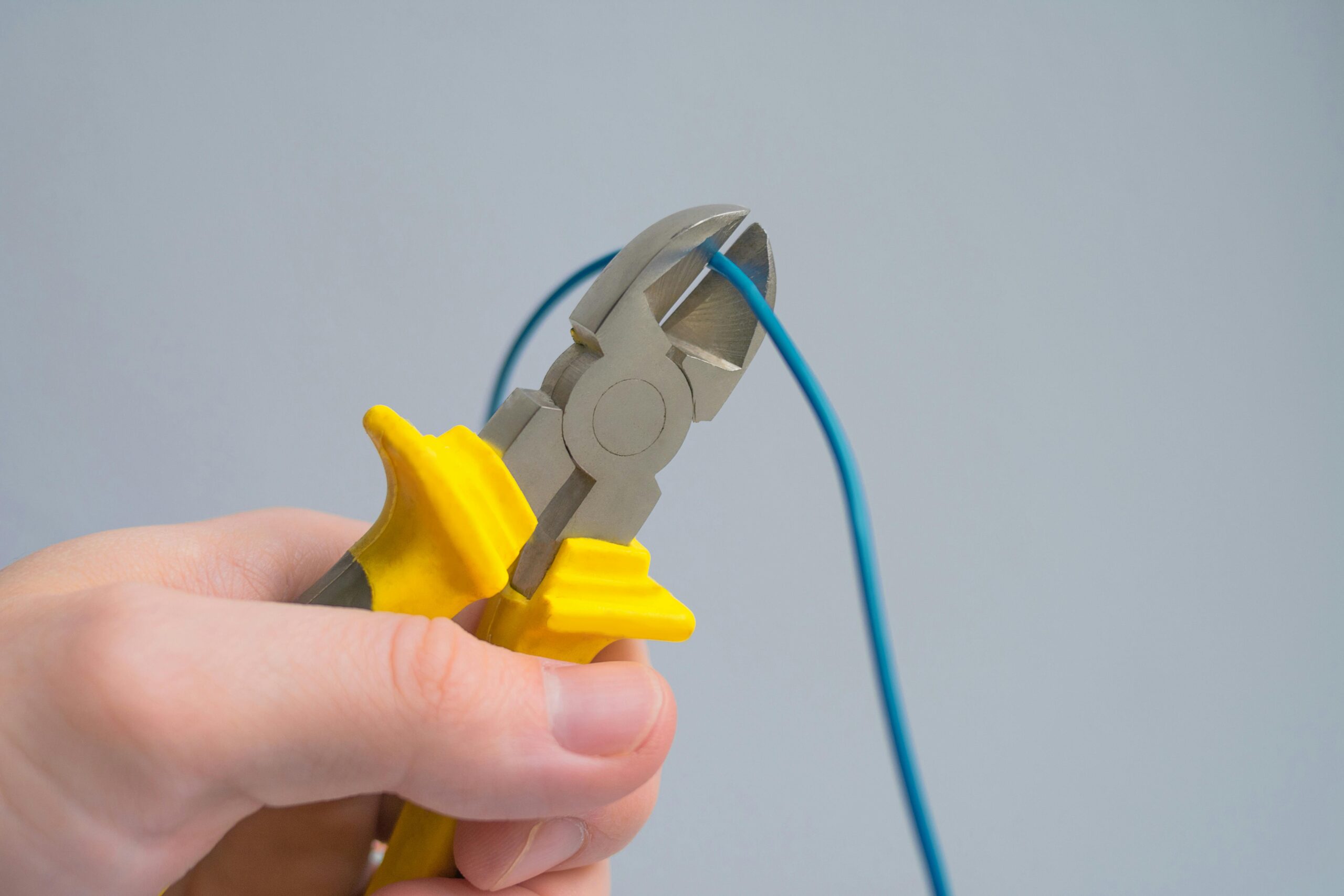 A close-up image of a hand using pliers to cut a blue wire against a gray background.